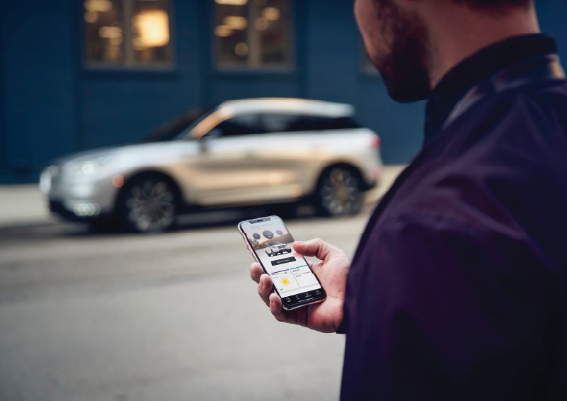 A man is holding his phone looking at the Lincoln Way App and we see his 2021 Lincoln Corsair in the background against a blue brick wall