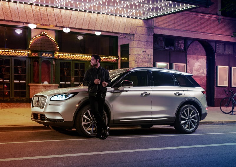A 2022 Lincoln Corsair SUV is parked outside a theater as the driver relaxes against the frame and lights illuminate the floating roofline and body | Empire Lincoln in Abingdon VA