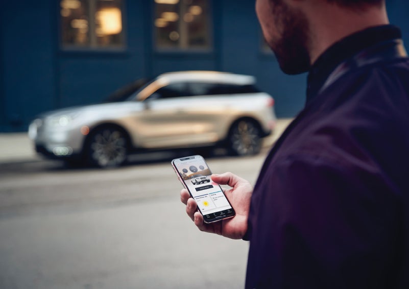 A man is holding his phone looking at the Lincoln Way™ App and we see his 2022 Lincoln Corsair SUV in the background against a blue brick wall | Empire Lincoln in Abingdon VA